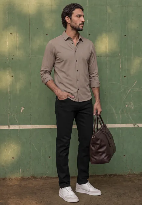 Een man in beige overhemd, zwarte broek en witte sneakers draagt een bruine tas voor een groene muur.