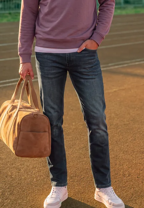 Person in Jeans, weißen Sneakern und rosa Sweatshirt hält braune Reisetasche auf Laufbahn.