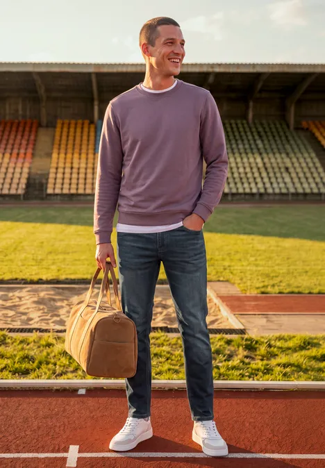 Ein Mann in lila Sweatshirt, Jeans und weißen Sneakern trägt eine Reisetasche auf der Stadion-Laufbahn.