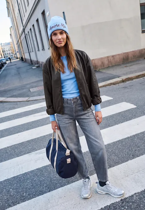 Junge Frau in Jeans, Bomberjacke und Mütze mit Tasche steht auf einem Zebrastreifen.