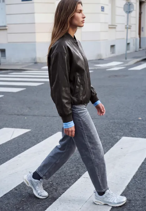 Junge Frau in Lederjacke, Jeans und Sneakers läuft über einen Zebrastreifen in der Stadt.