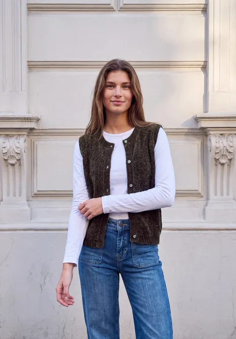 Frau lehnt lächelnd an einer Fassade, trägt weißes Shirt, Weste und Jeans.