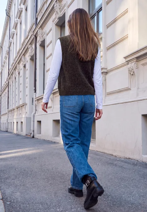 Eine Person mit blauer Jeans, schwarzer Weste und weißem Longsleeve spaziert an einem weißen Gebäude vorbei.