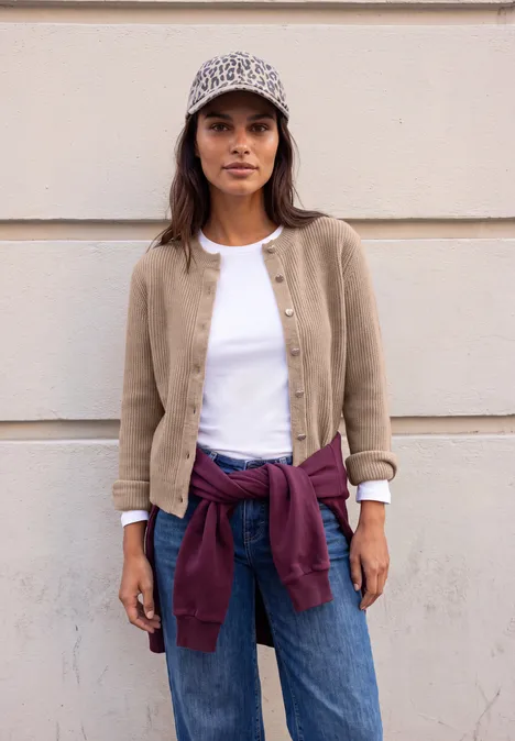 Eine junge Frau vor heller Wand, trägt beige Jacke, weißes Shirt, Jeans, lila Pullover um die Taille und ein gemustertes Cap.
