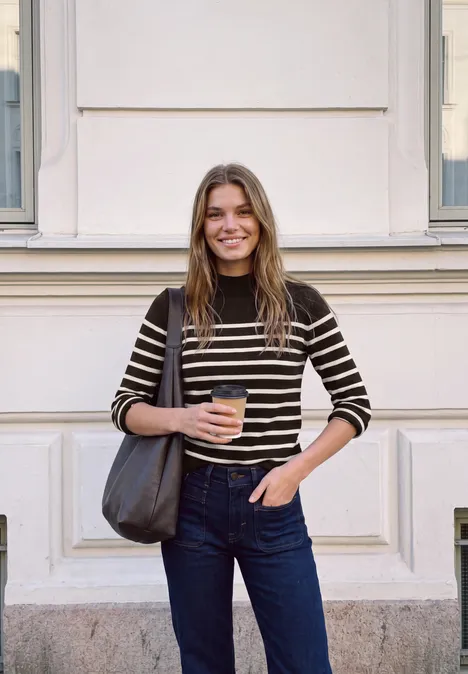 Eine junge Frau steht vor einer Fassade, hält einen Coffee-to-go und trägt Ringelshirt und Jeans.