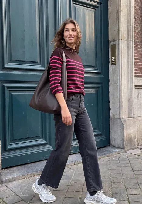 Eine Person in gestreiftem Pullover, dunkler Hose und Schultertasche steht vor einer Tür.