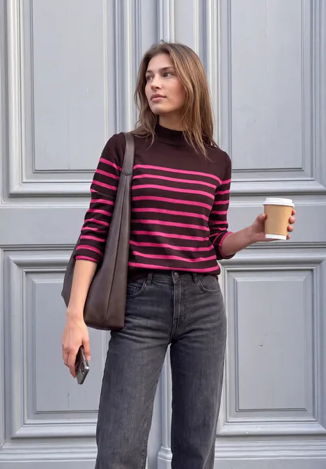 Eine junge Frau steht vor einer Tür, hält Kaffee und Smartphone, trägt gestreiftes Shirt und Jeans.