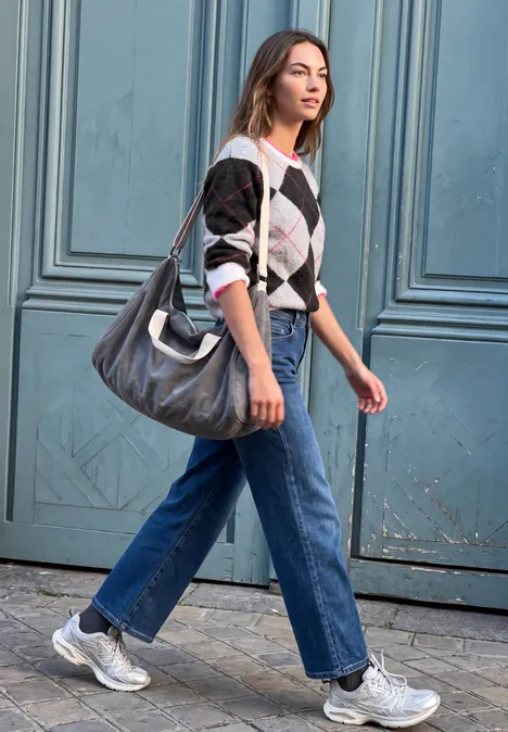 Frau in Jeans, Sneakers und Pullover mit großer Tasche geht an einer Haustür vorbei.