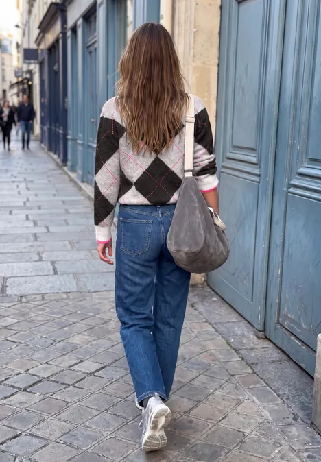 Frau in Jeans, Rautenmuster-Pullover und weißen Sneakers läuft auf Kopfsteinpflaster.