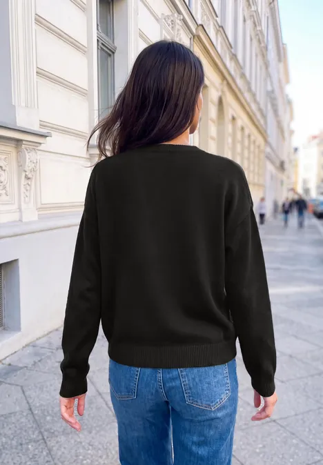 Pullover mit großem Wording Artwork Black Frau von hinten in schwarzem Pullover und Jeans, stehend in einer europäischen Altstadtstraße.