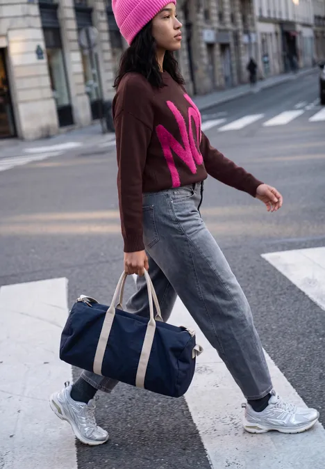 Pull à slogan russet red Femme en bonnet rose et pull traverse la rue en ville, portant un sac de voyage bleu et des baskets.