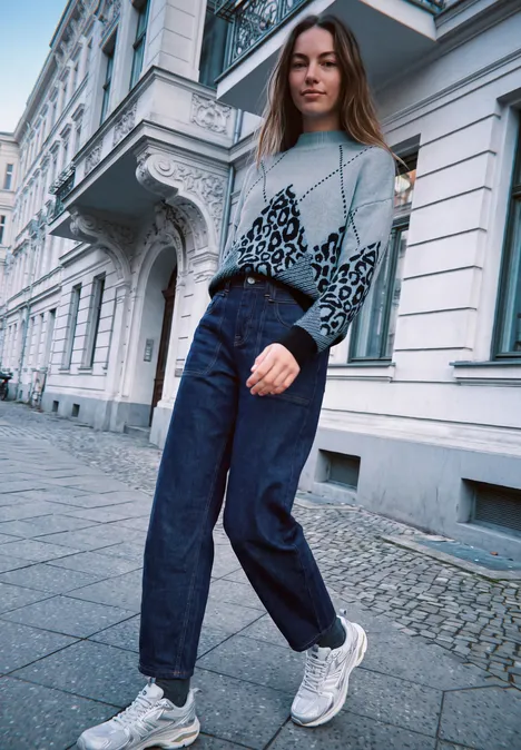 Jonge vrouw in gebreide trui, wijde broek en sneakers loopt langs een stijlvolle stadsgevel.