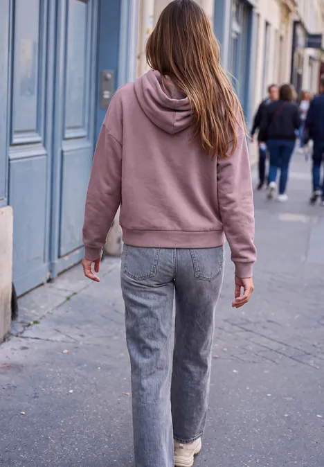 Eine Person in rosa Kapuzenpullover und grauen Hosen geht auf einem Bürgersteig in der Stadt.