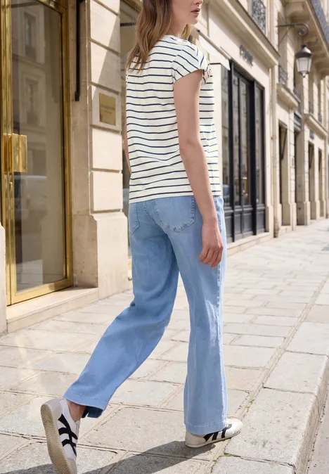 Frau in gestreiftem T-Shirt, blauer Jeans und Sneakern spaziert in einer schicken Fußgängerzone.