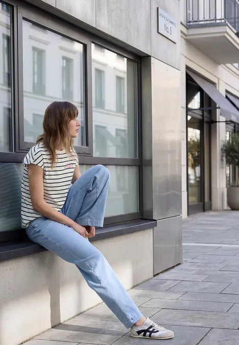 Eine junge Frau sitzt lässig auf einer Fensterbank an einer Hausfassade in der Stadt, trägt gestreiftes Shirt und Jeans.