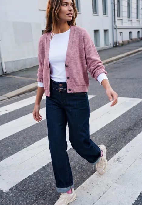 Eine Frau spaziert auf einem Zebrastreifen, trägt weißes Shirt, rosa Cardigan, weite Jeans und Sneaker.