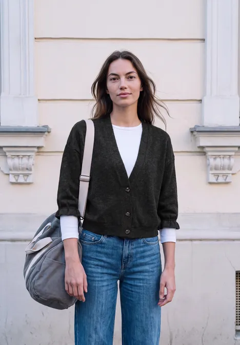 Jeune femme en cardigan, t-shirt blanc, jean et sac, debout devant une façade claire.