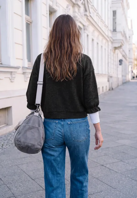 Une femme en jean bleu et pull noir marche dans la rue, portant un sac gris.