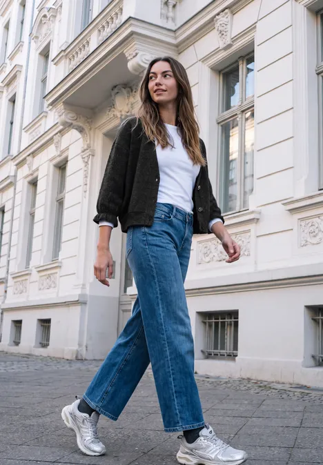 Jeune femme en jean, t-shirt blanc et baskets argentées marche devant un élégant immeuble ancien.