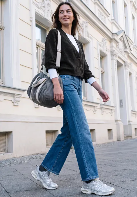 Jeune femme en jean, veste noire et baskets argentées marche en souriant devant une façade claire.