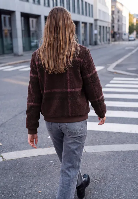 Eine Person in brauner Jacke und Jeans geht auf dem Zebrastreifen einer urbanen Straße.