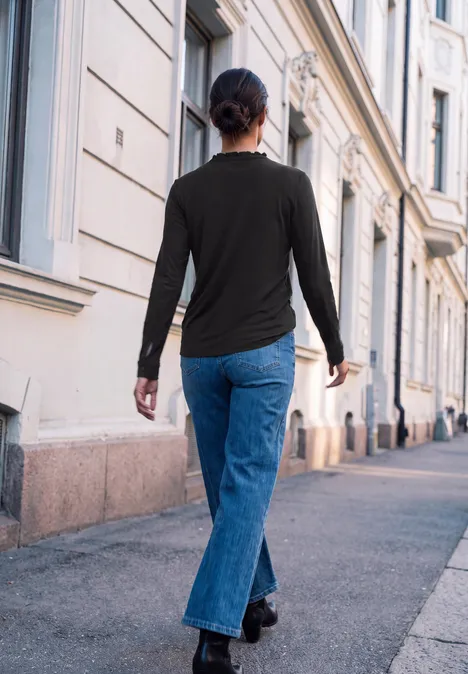 Shirt met lange mouwen, ronde hals en ruches Black Eine Frau geht von hinten auf einem Gehweg entlang, flankiert von klassischen Gebäuden.
