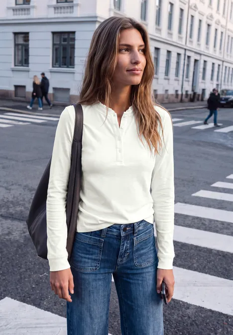 Shirt met lange mouwen, ronde hals en ruches off white Junge Frau in weißem Langarmshirt und Jeans geht über eine Straßenkreuzung, trägt Tasche und Handy.