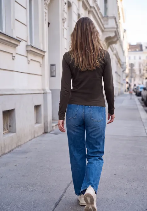 Shirt met lange mouwen, ronde hals en ruches charred brown Frau in lässiger Kleidung läuft auf einem städtischen Gehweg mit Häuserfassaden im Hintergrund.
