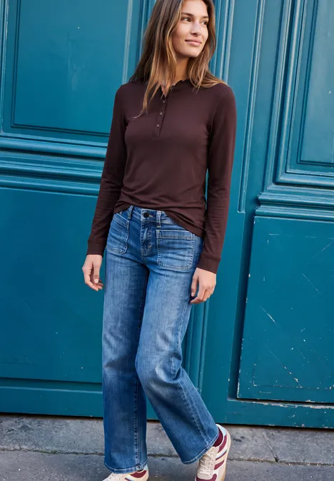Shirt met lange mouwen, ronde hals en ruches russet red Frau steht vor einer blauen Tür, trägt braunes Oberteil, Jeans und Sneaker, lächelt und schaut zur Seite.