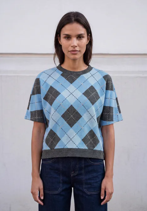Shirt met gebreide look en argylepatroon loom blue Frau trägt kurzes blau-graues Rauten-Pullover-Shirt und dunkle Jeans vor neutralem Hintergrund.
