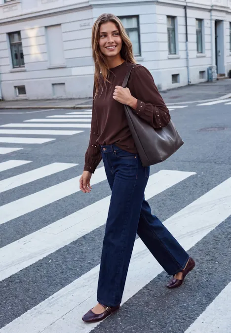 Chemisier à manches longues avec des rivets et un col smocké russet red Une femme en haut marron et jean traverse en souriant un passage piéton en ville avec un sac.