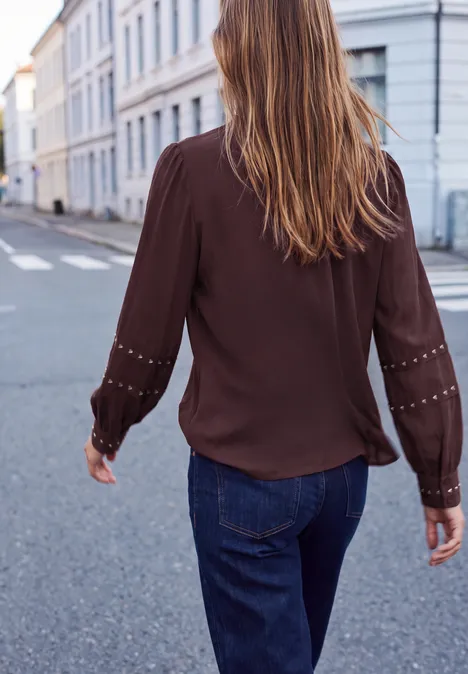 Chemisier à manches longues avec des rivets et un col smocké russet red Femme de dos en chemisier marron et jean marche dans une rue calme bordée de maisons claires.