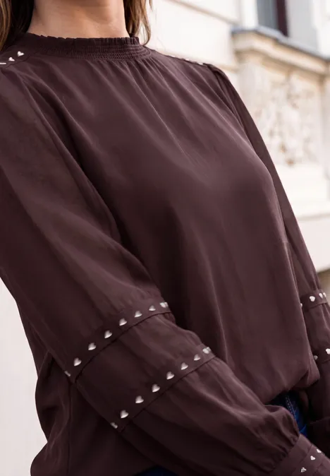 Chemisier à manches longues avec des rivets et un col smocké russet red Blouse brun foncé à manches longues avec ornements clous aux poignets, portée devant un bâtiment.