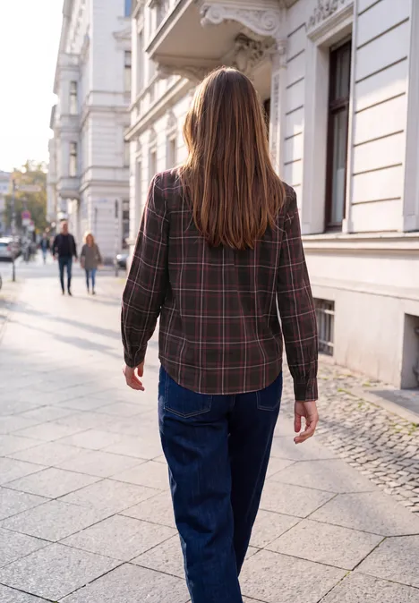 Une femme en chemise à carreaux et jeans marche dans une rue urbaine bordée d'immeubles.