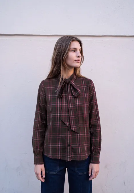 Femme portant un chemisier marron à carreaux avec un nœud au col, debout devant un mur clair, regard de côté.