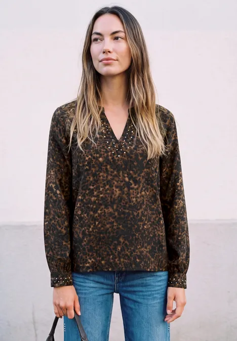 Tunique à col fendu et détails rivetés charred brown Femme en chemisier foncé à motif et jean bleu devant un mur clair.