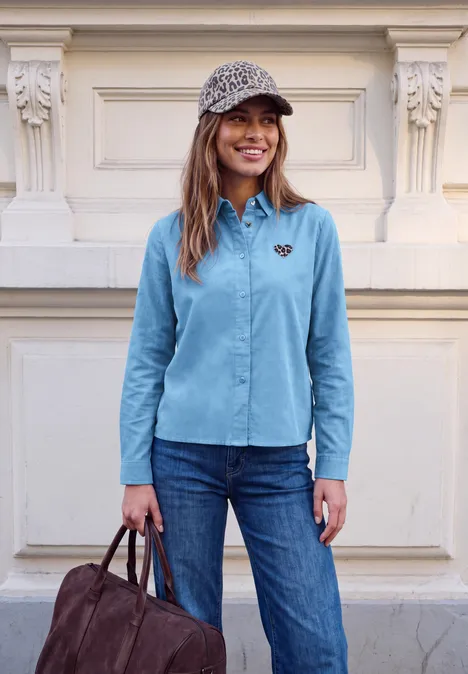 Junge Frau vor cremefarbener Haustür, trägt hellblaues Hemd, Jeans, gemustertes Cap, hält braune Tasche, lächelt.