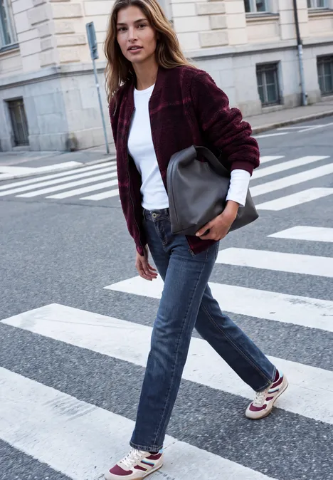 Eine junge Frau in Jeans, weißem Shirt und bordeauxrotem Cardigan überquert eine städtische Straße mit Handtasche.