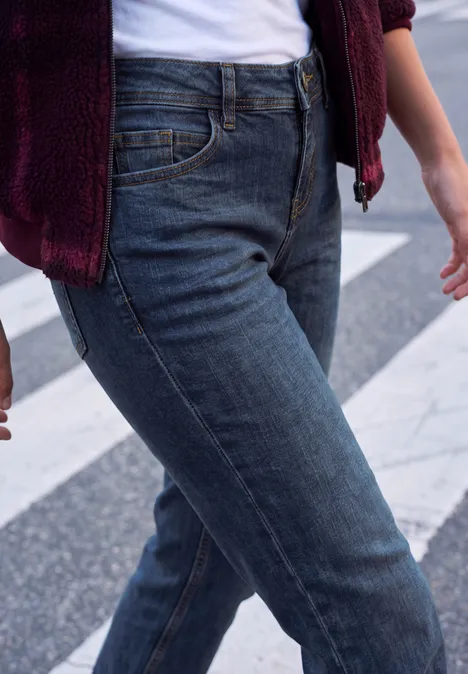 Nahaufnahme einer Person in blauen Jeans und burgunderrotem Oberteil auf einem Zebrastreifen.
