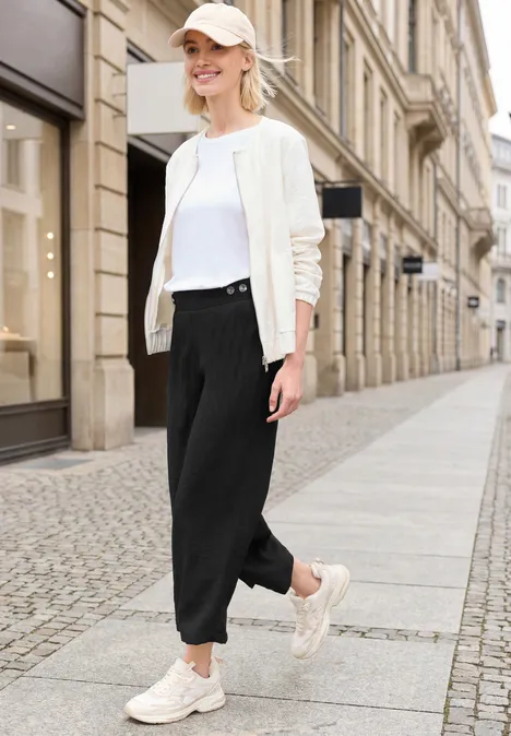 Modische Frau in heller Jacke, weißem T-Shirt und schwarzen Jogginghosen spaziert auf städtischem Gehweg.