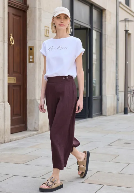 Person in weißem T-Shirt und weinroter Hose mit Sandalen spaziert auf einem Gehweg.