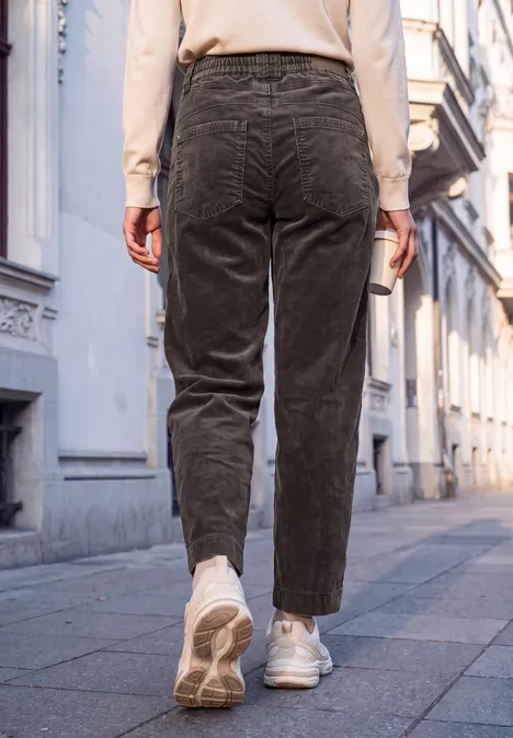 Eine Person in Cordhosen und weißen Sneakern geht auf einem Gehweg zwischen Altbauten.