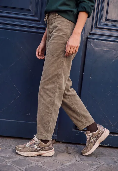 Eine Person trägt beige Cordhosen und braune Sneaker vor einer dunklen Tür
