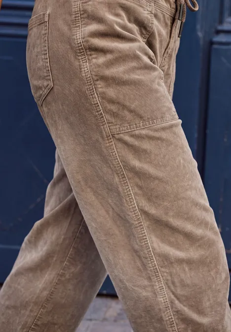 Braune Cordhose an Personenbeinen, seitliche Nahaufnahme vor dunklem Hintergrund.