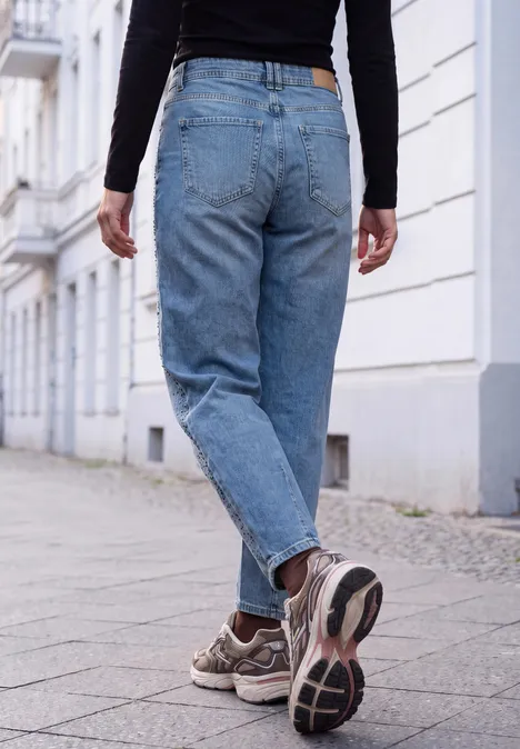 Widok od tyłu osoby w niebieskich dżinsach i sneakersach, idącej chodnikiem w cichej, miejskiej okolicy.