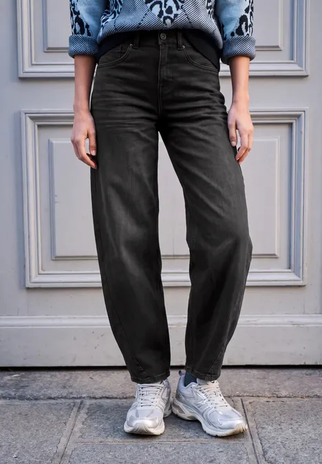 Person steht vor einer grauen Tür, trägt weite schwarze Hose und weiße Sneaker.