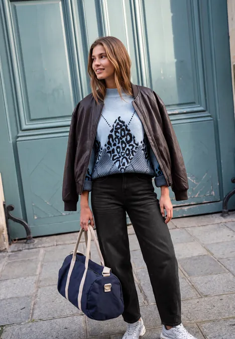 Junge Frau mit Lederjacke, gemustertem Pullover, schwarzer Hose und Reisetasche vor blauem Tor.