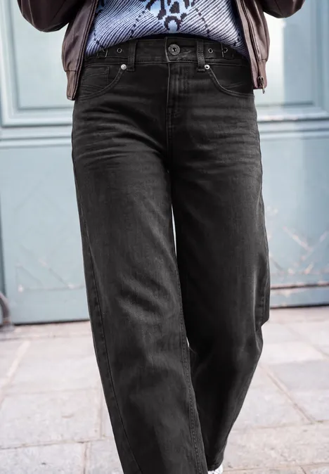 Schwarze, gerade geschnittene Jeans an einer Person, kombiniert mit gemustertem Hemd vor blauer Tür.