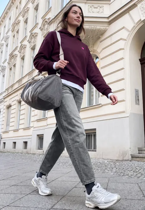 Relaxed Barrel Leg Jeans im Loose Fit grey authentic washed Eine junge Frau in Kapuzenpullover, Jeans und Sneakern trägt eine Sporttasche und geht eine Straße entlang.