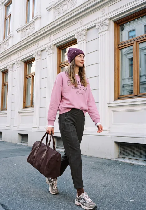 Mid Waist Barrel Leg Hose im Loose Fit inked grey Junge Frau in pinkem Hoodie und Mütze spaziert mit Tasche vor klassischer Fassade.